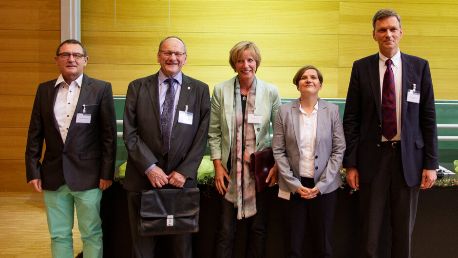 Fünf Personen in formeller Kleidung stehen für ein Gruppenfoto in einer Reihe nebeneinander in einem Hörsaal.
