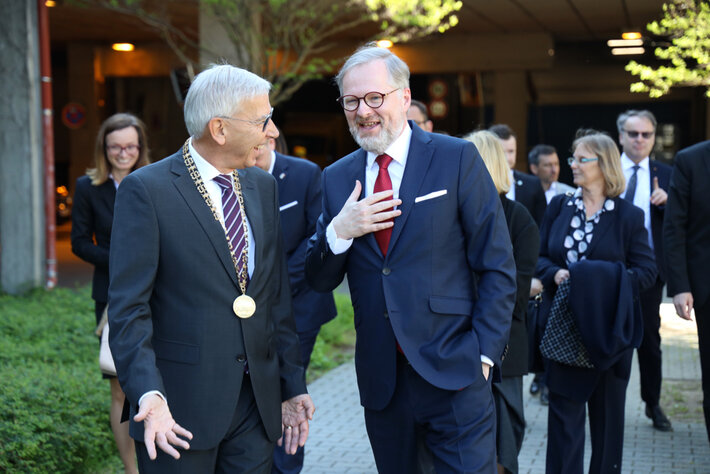 Zwei Männer unterhalten sich lächelnd miteinander. Die Person auf der linken Seite trägt eine Amtskette. Im Hintergrund sind mehrere, formell gekleidete Personen zu sehen. Alle Personen sind in Bewegung, sie scheinen zu gehen. (öffnet Vergrößerung des Bildes)