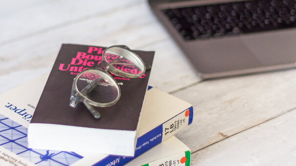 Ein Stapel Bücher, eine Brille und ein offener Laptop liegen auf einem Schreibtisch.