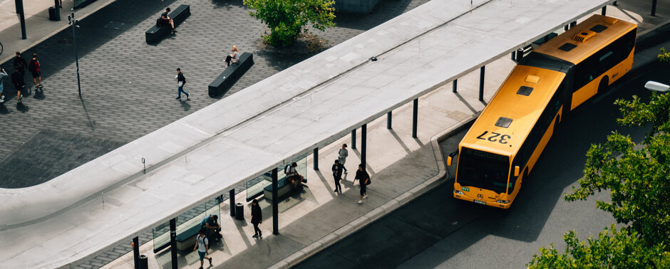 Eine große Omnibushaltestelle aus der Vogelperspektive. Man sieht Fahrgäste auf dem Bussteig; ein gelber Gelenkbus verlässt gerade eine der Haltebuchten.
