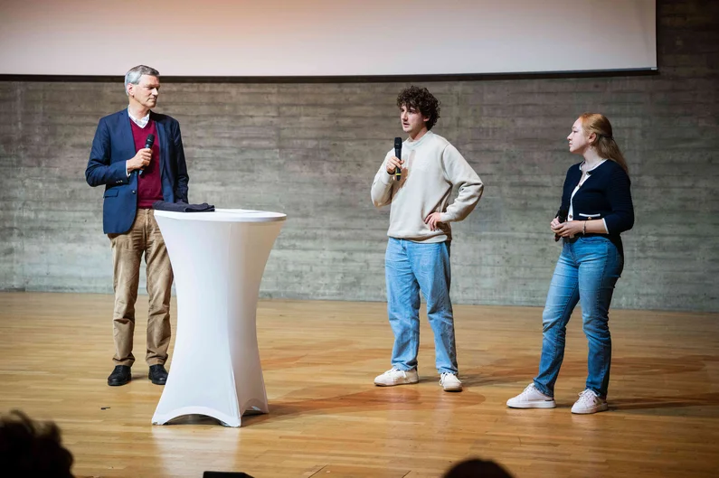 Drei Personen stehen auf einer Bühne um ein weißes Rednerpult und halten Mikrofone. Im Rahmen einer Gesprächsrunde stellen sie studentische Vertretungen vor. (öffnet Vergrößerung des Bildes)