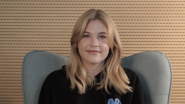 Porträt einer jungen Frau mit langen blonden Haaren, die in einem grauen Sessel vor einem braunen Hintergrund sitzt.