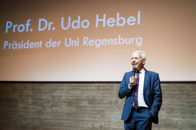 Eine Person in blauem Anzug steht mit einem Mikrofon auf einer Bühne. Auf der Leinwand im Hintergrund sind die Worte „Prof. Dr. Udo Hebel“ und „Präsident der Uni Regensburg“ zu sehen. (öffnet Vergrößerung des Bildes)