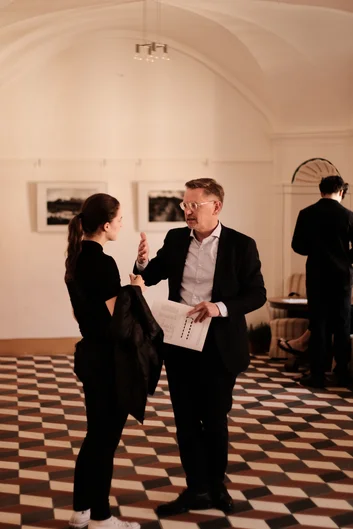 Ein Mann im Anzug spricht mit einer Frau in legerer Kleidung in einem elegant dekorierten Raum mit schwarz-weiß kariertem Boden. Er hält ein Blatt Papier in der Hand, während sie aufmerksam zuhört. Im Hintergrund unterhalten sich zwei weitere Personen vor gerahmten Fotografien an der Wand. (öffnet Vergrößerung des Bildes)