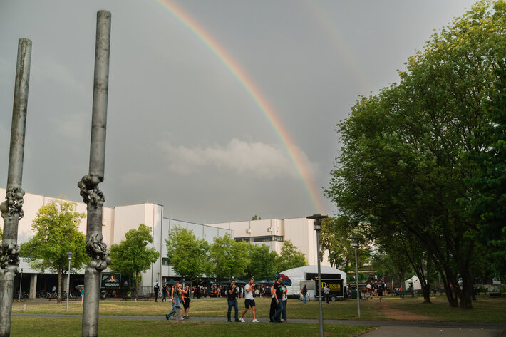 Ein Regenbogen über dem PT-Gebäude der Universität Regensburg. Zu sehen sind außerdem zwei Stehlen - Teil eines Kunstwerkes vor dem Vielberth-Gebäude -, einzelne Personen und Bäume (öffnet Vergrößerung des Bildes)