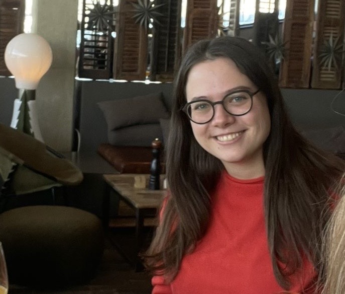 Eine junge Frau mit Brille und braunen Haaren lächelt in die Kamera. Sie sitzt in einem öffentlichen Raum ähnlich einer Hotellobby. Im Hintergrund sieht man Sofas und einen Raumtrenner.