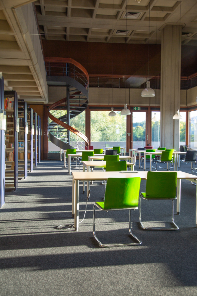 Blick in den Allgemeinen Lesesaal: Grüne Stühle vor weißen Tischen, am linken Rand sind Bücherregale zu erkennen, im Hintergrund eine Wendeltreppe und große Fensterfronten, die Ausblick auf Bäume und Rasenflächen geben.