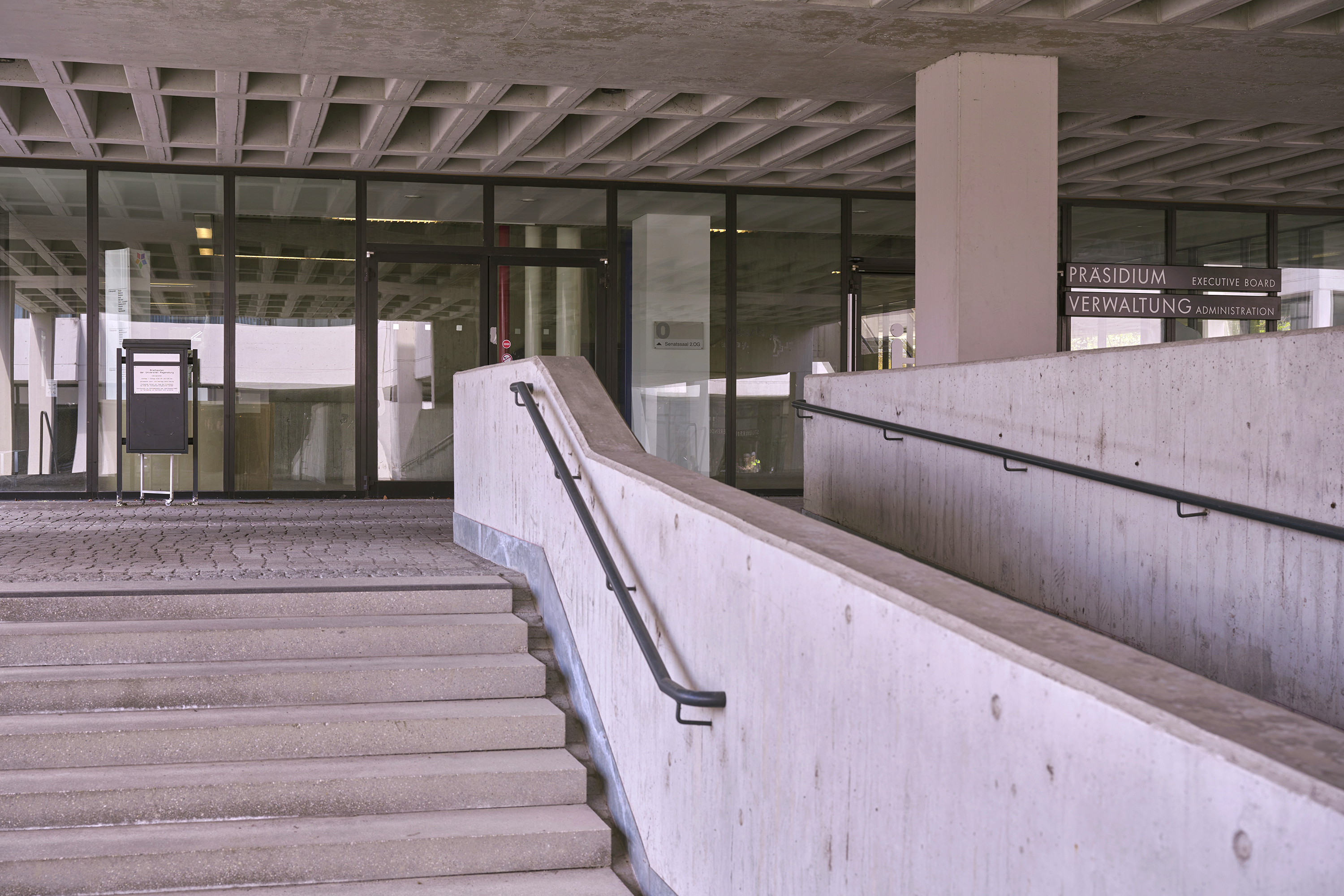 Eingangsbereich eines Gebäudekomplexes in Sichtbetonbauweise; vor einer Glasfront ist ein großformatiger Briefkasten zu sehen. Im Bildvordergrund sind Treppenstufen zu sehen. (öffnet Vergrößerung des Bildes)