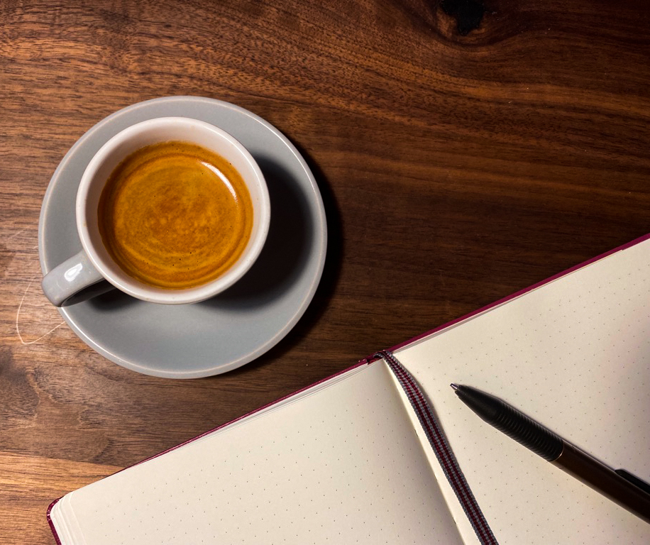 Eine Tasse Kaffee steht auf einer Oberfläche aus Holz, daneben liegt ein aufgeschlagenes Notizbuch mit Lesebändchen und Kugelschreiber.