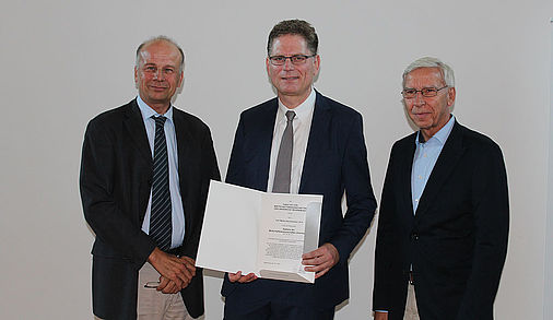 Drei Männer in dunkeln Anzügen stehen nebeneinander. Der Mann in der Mitte hält eine aufgeklappten Urkundenmappe in den Händen.