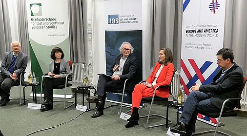 Fünf Personen in formeller Kleidung sitzen auf in einem Halbkreis angeordneten Stühlen. Der in der Mitte sitzende ältere Mann mit hellgrauen Haaren und schwarz umrandeter Brille hält ein Mikrofon in der Hand und schaut zur Sitznachbarin zu seiner Linken.