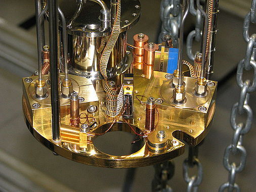 Nahaufnahme eines komplexen mechanischen oder elektronischen Geräts, vermutlich Teil eines wissenschaftlichen Instruments. Sichtbar sind gold- und kupferfarbene Metallkomponenten, Drähte, Schrauben sowie mehrere zylindrische Strukturen und Ketten im Hintergrund.