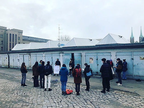 Eine Gruppe von circa 15 Personen in winterlicher Straßenkleidung steht in einem losen Kreis in einer Art Garagenhof. Manche tragen eine FFP2-Maske. Ein Mann hält ein Plakat in den Händen.