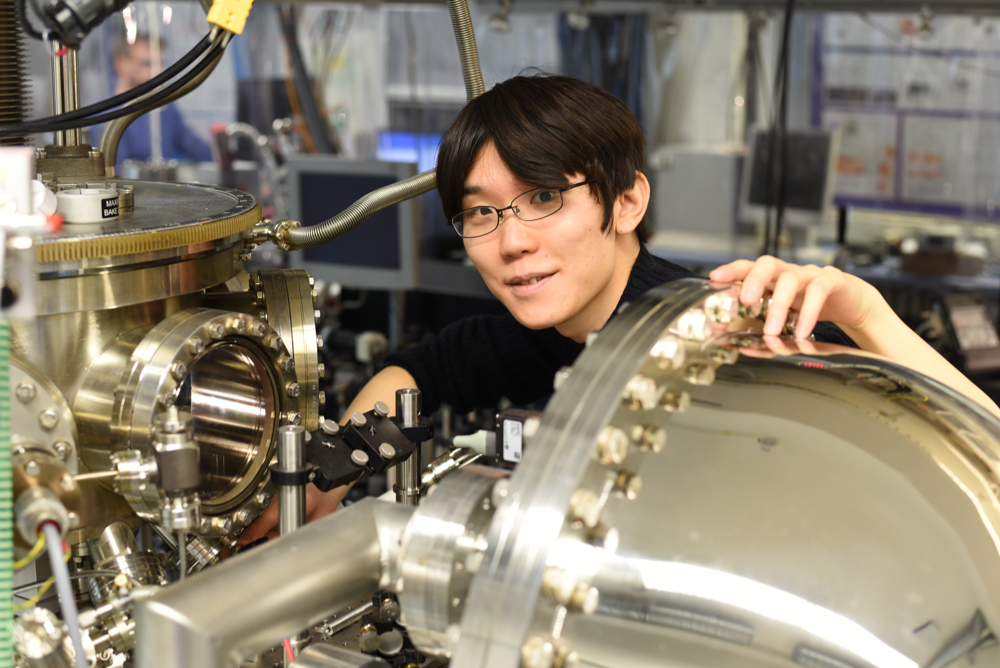 Person working with complex scientific equipment in a laboratory. The setup includes metal components, tubes, valves, and measuring instruments. Computer monitors and other electronic devices are visible in the background. 