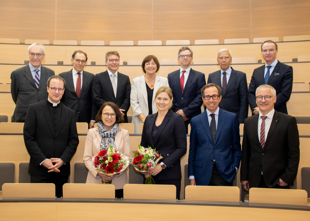 Ein Gruppenbild mit 12 Personen in formeller Kleidung, die sich auf zwei Reihen in einer Hörsaalbestuhlung verteilen. Zwei Frauen in der vorderen Reihe halten Blumensträuße in den Händen.