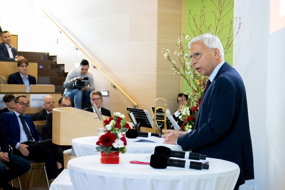 Ein Mann mit weißen Haaren im dunklen Anzug steht an mit weißen Hussen überzogenen Stehtischen und spricht Richtung Publikum. Im Hintergund sind Teilnehmende der Veranstaltung auf Hörsaalbestuhlung zu erkennen. 