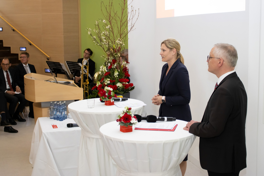 Zwei Personen in dunklen Anzügen stehen an mit weißen Hussen überzogenen Stehtischen. Auf den Tischen stehen Blumengestecke und liegen Dokumente und Mirkofone. Im Hintergrund sitzen Musiker, die auf ihren Auftritt warten, und Gäste der Veranstaltung. 