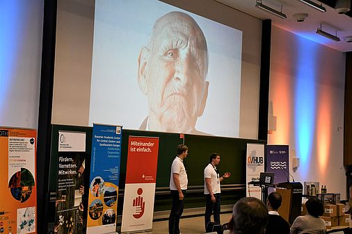 Der vordere Bereich eines Hörsaals, in dem mehrere Roll-Ups aufgebaut sind. Zwei Männer in weißen T-Shirts halten einen Vortrag. Im Hintergrund wird das Bild eines alten Mannes mit mürrischem Gesichtsausdruck an die Wand projeziert.