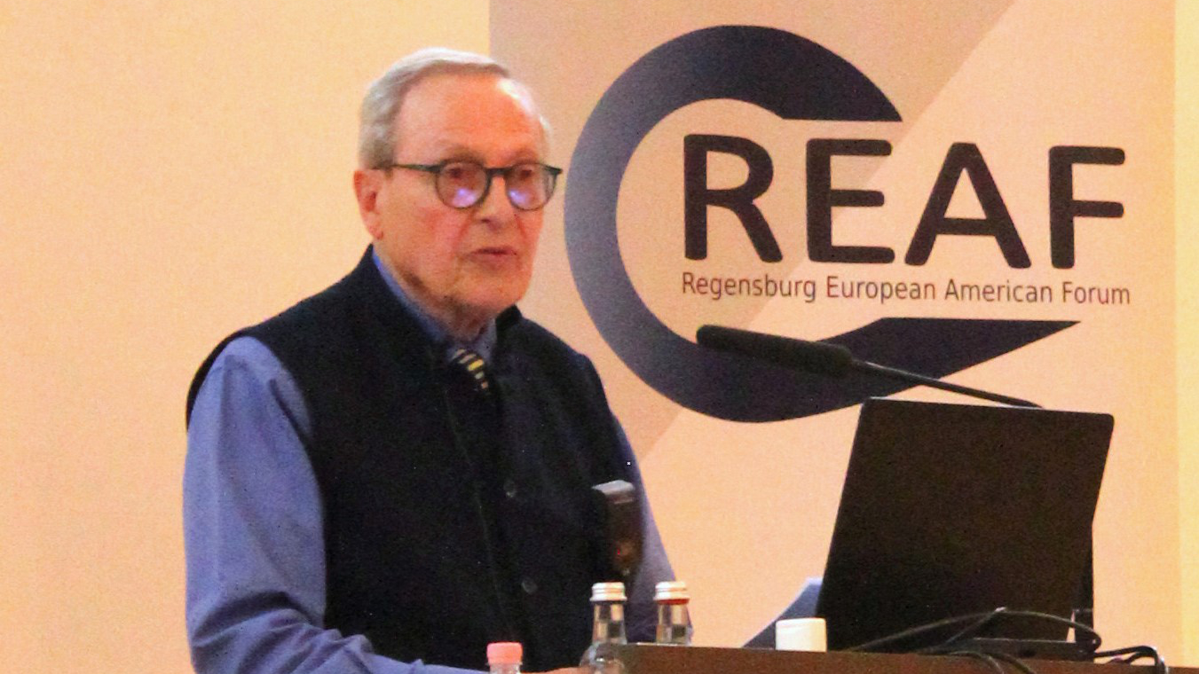 Ein älterer Mann in blauem Hemd und schwarzer, hochgeschlossener Weste steht an einem Redner-Pult. Im Hintergrund, an der Wand, steht der Schriftzug: "REAF Regensburg European American Forum".