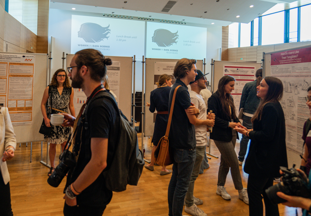 Mehrere Personen stehen in einem Veranstaltungsraum, der mit Palaktwänden strukturiert ist. Manche unterhalten sich in Zweiergruppen, manche Personen studieren die wissenschaftlichen Plakate. 