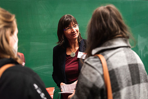Eine Frau mit Brille steht vor einer grünen Tafel und spricht mit zwei Personen.