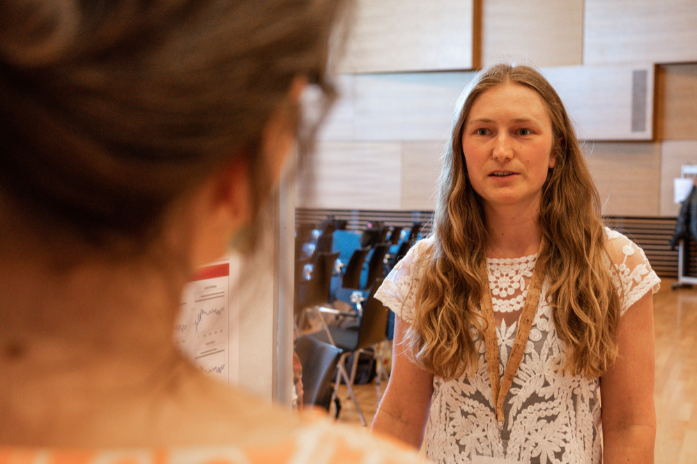 A woman with long blonde hair is talking to another person, of whom only the back of the head is vaguely visible.
