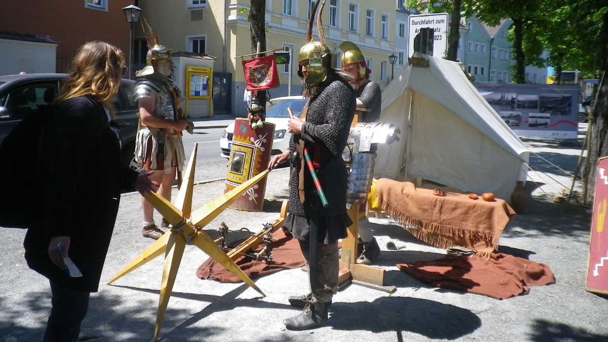 Auf einem breiten Bürgersteig gegenüber von historischen Wohnhäusern wurde ein Zelt aufgebaut. Davor stehen drei Männer in römischen Legionärsrüstungen. An einem Baum lehnt ein Schutzschild, darüber hängt ein rotes Banner. Einer der Männer ist im Gespräch mit einer Passantin.