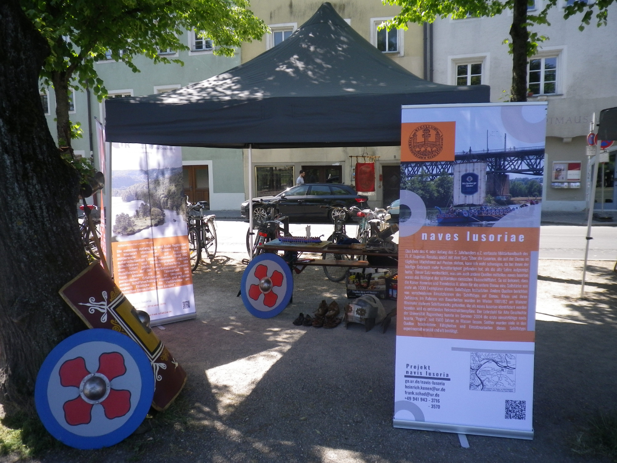 Ein Pavillion überdacht einen Biertisch, auf und vor dem Teile einer römischen Rüstung liegen. Daneben stehen Banner mit Informationen zu "naves lusoriae". 
