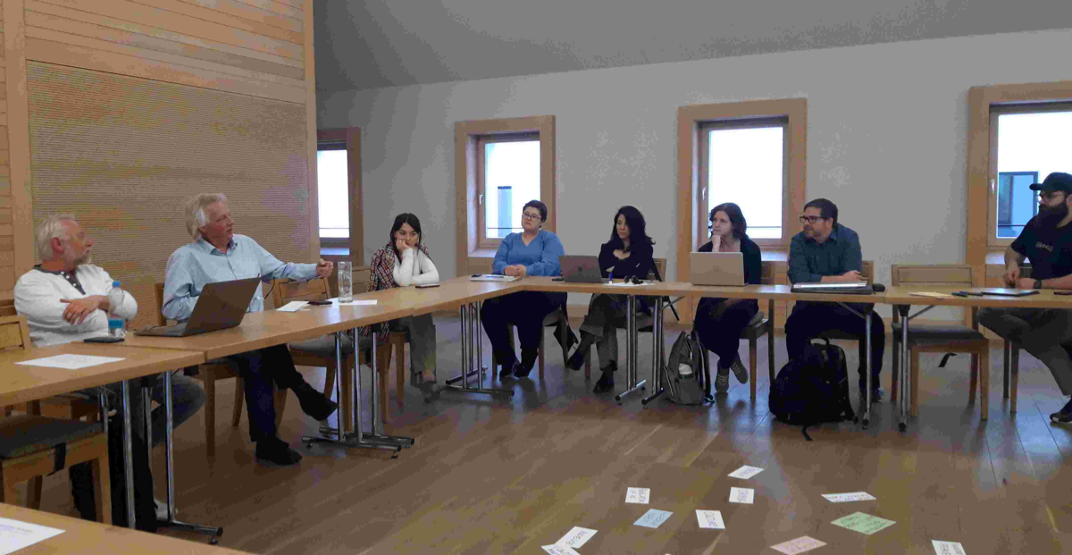 Mehrere Personen sitzen an einem Konferenztisch, der über Eck steht. 