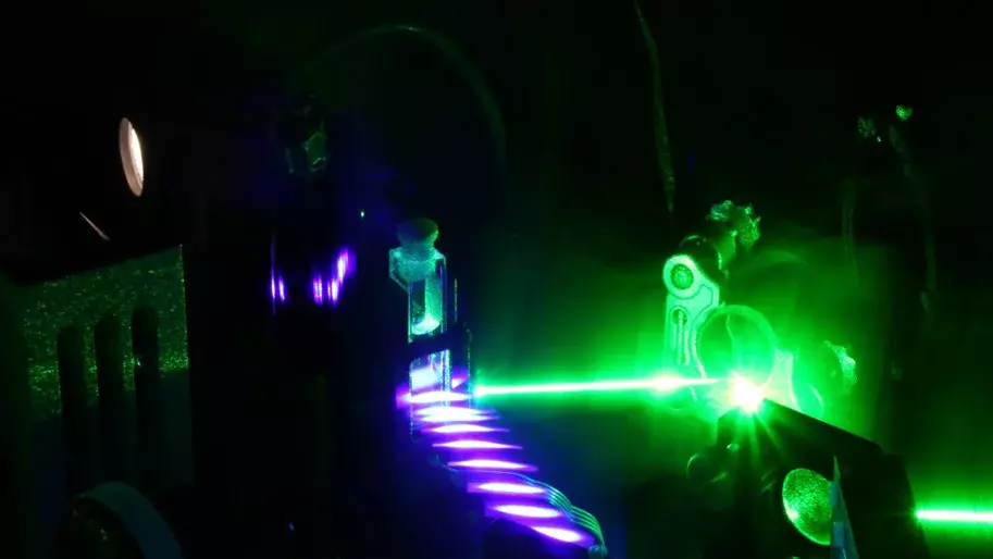 Vor einem schwarzen Hintergrund lassen sich schattenhaft technische Gerätschaften in einer Versuchsanordnung erahnen. Links ist eine Reihe langgezogener Ellipsen mit nahezu weißem Lichtkern umgeben von jeweils einem violetten Lichtkran zu sehen. Ein dünner, grün umrandeter Lichtstrahl verbindet den Anfang dieser Reihe mit einem Gerät auf der rechten Seite. 