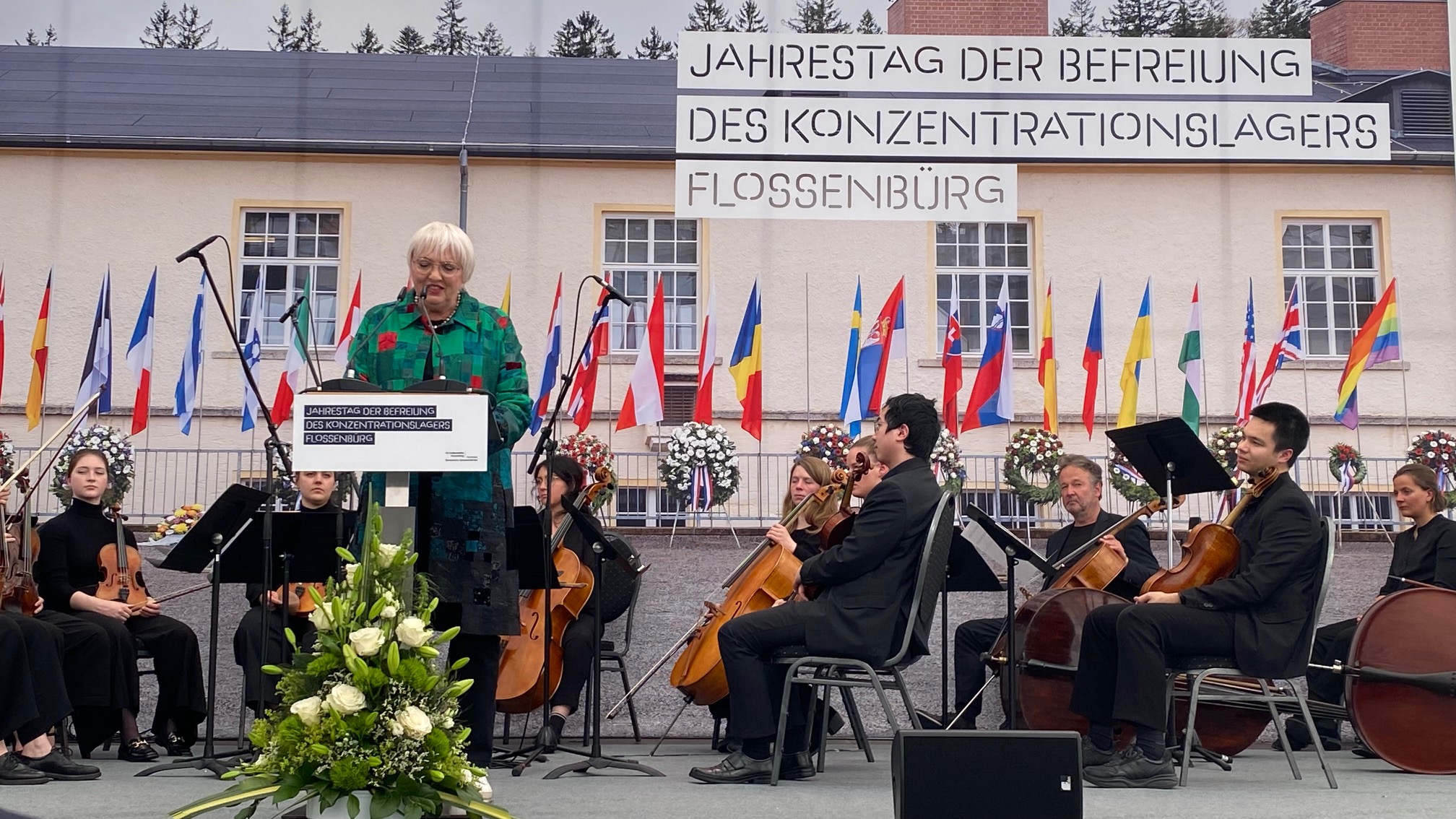 Eine ältere Frau steht an einem Rednerpult auf einer Bühne und spricht bei einer Gedenkveranstaltung. Hinter ihr sitzen Musikerinnen und Musiker eines Streichorchesters. Im Hintergrund hängen viele internationale Flaggen an einem Gebäude. Über dem Gebäude ist ein großes Banner mit dem Text: „Jahrestag der Befreiung des Konzentrationslagers Flossenbürg“ angebracht. Vor dem Rednerpult steht ein großer Blumenkranz. Die Veranstaltung findet im Freien statt. (öffnet Vergrößerung des Bildes)