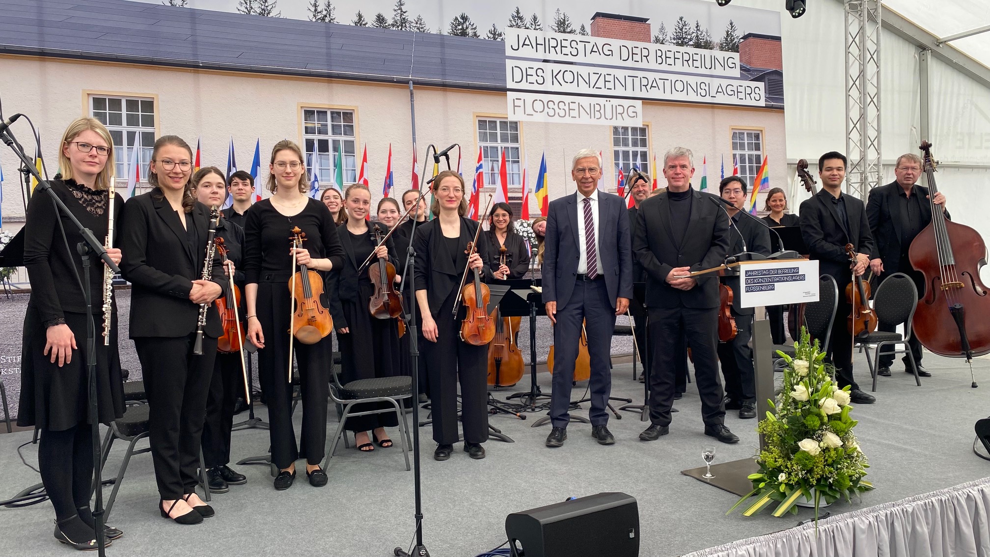 ine Gruppe von Musikerinnen und Musikern steht auf einer Bühne und blickt lächelnd in die Kamera. Sie halten verschiedene Streich- und Blasinstrumente in den Händen. Neben ihnen stehen zwei Männer im Anzug bzw. in dunkler Kleidung. Im Hintergrund sind internationale Flaggen und ein Gebäude zu sehen. Über dem Gebäude hängt ein großes Banner mit der Aufschrift: „Jahrestag der Befreiung des Konzentrationslagers Flossenbürg“. Vor der Bühne steht ein Blumenarrangement. Die Aufnahme wurde im Rahmen einer Gedenkveranstaltung gemacht. (öffnet Vergrößerung des Bildes)