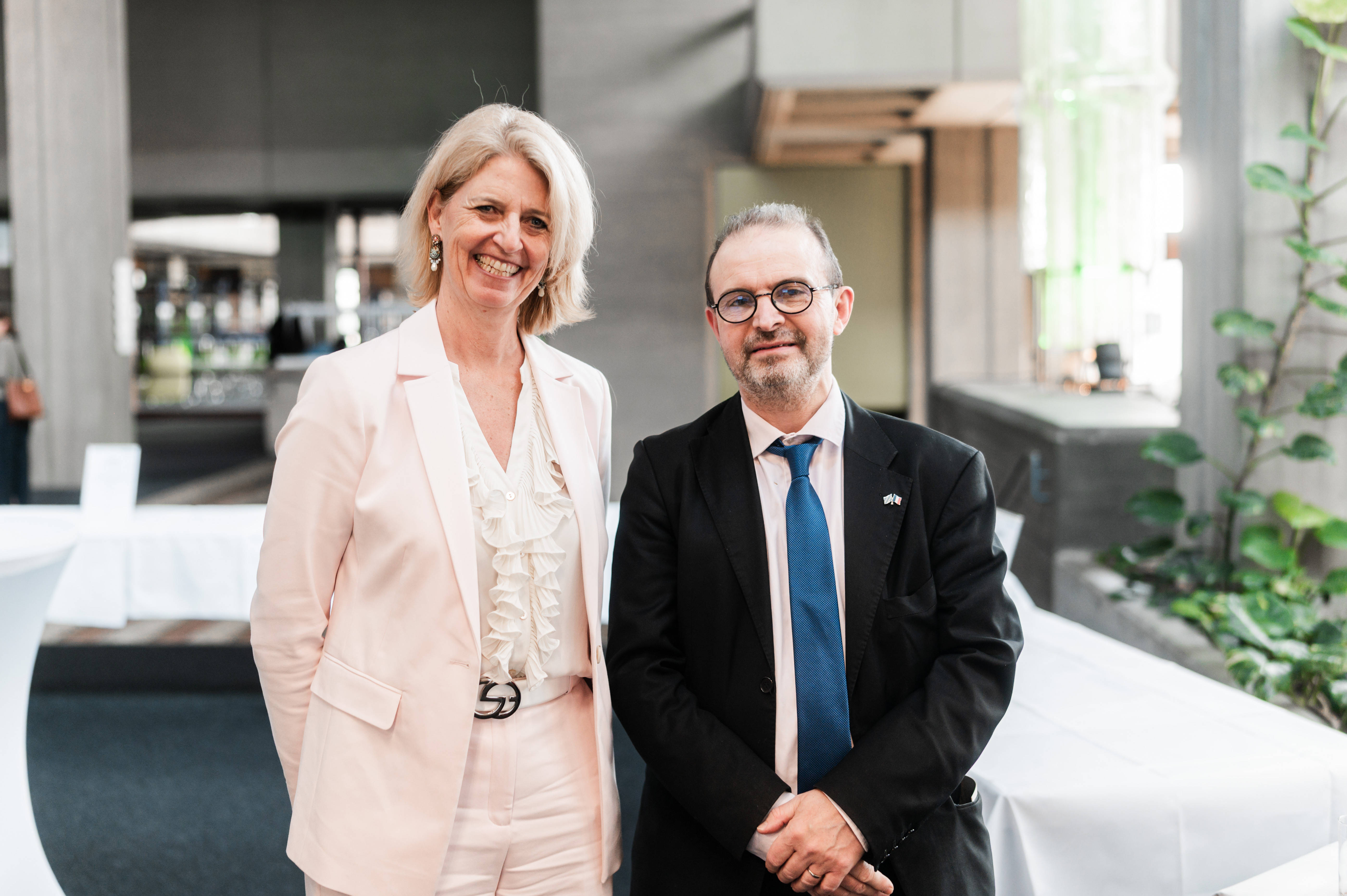 Eine Frau im weißen Hosenanzug und ein Mann im schwarzen Anzug stehen nebeneinander und lächeln in die Kamera. (öffnet Vergrößerung des Bildes)