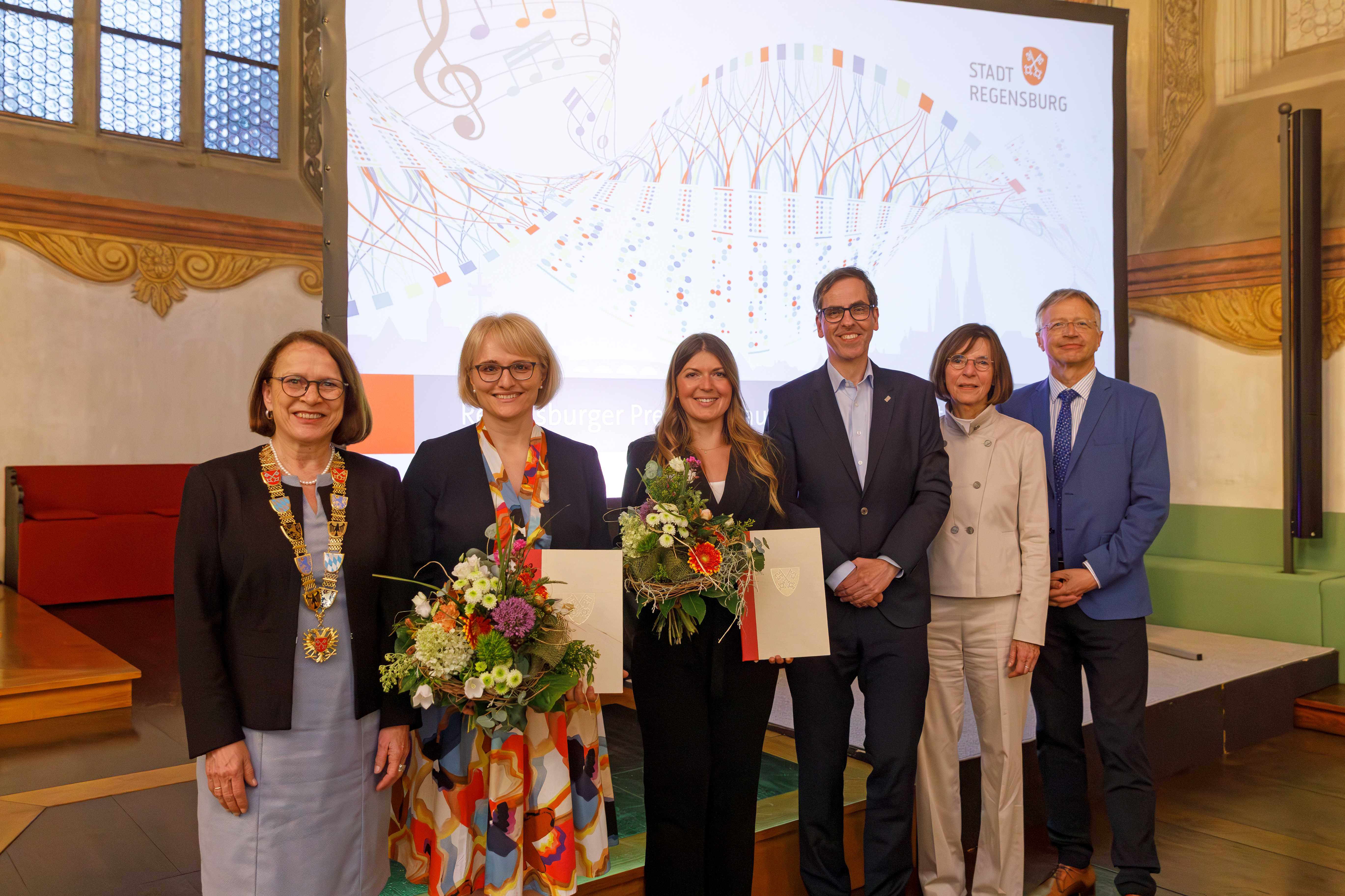 Das Gruppenfoto zeigt sechs Personen. Von links nach rechts: Die erste Frau trägt ein hellblaues Kleid und einen schwarzen Blazer, darüber trägt sie die Amtskette der Stadt Regensburg, daneben steht eine Frau mit einem bunten Kleid und einem schwarzen Blazer. In der Hand hält sie einen bunten Blumenstrauß und eine Urkunde. Dann kommt eine Frau im schwarzen Anzug, sie hält einen Bluemstrauß und eine Urkunde. Daneben stehen ein Mann im schwarzen Anzug, eine Frau im weißen Anzug und ein Mann im blauen Anzug. (öffnet Vergrößerung des Bildes)