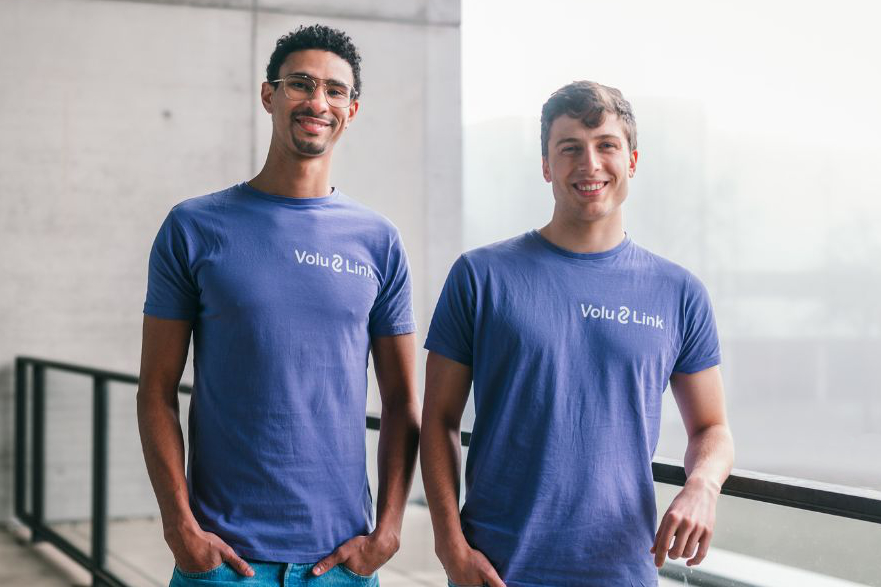 Zwei junge Männer in blauen T-Shirts mit dem Schriftzug "Volu Link" stehen an einer Balkonbrüstung und blicken lächelnd in die Kamera