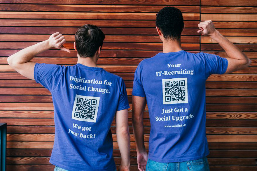 Zwei Personen stehen mit dem Rücken zum Betrachter vor einer holzvertäfelten Wand. Den jeweils äußeren Arm haben sie erhoben, der Ellbogen ist jeweils abgewinkelt und mit dem jeweiligen Daumen zeigen sie auf ihre T-Shirts. Dort sind Hinweise auf "VoluLink" und QR-Codes aufgedruckt.