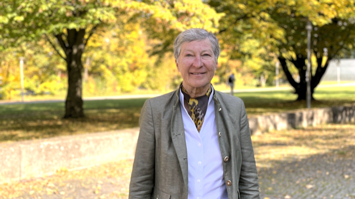 Eine Frau mit kurzen grauen Haaren blickt den Betrachter an. Im Hintergrund sind herbstliche gefärbte Laubbäume und eine Grünfläche zu erkennen.