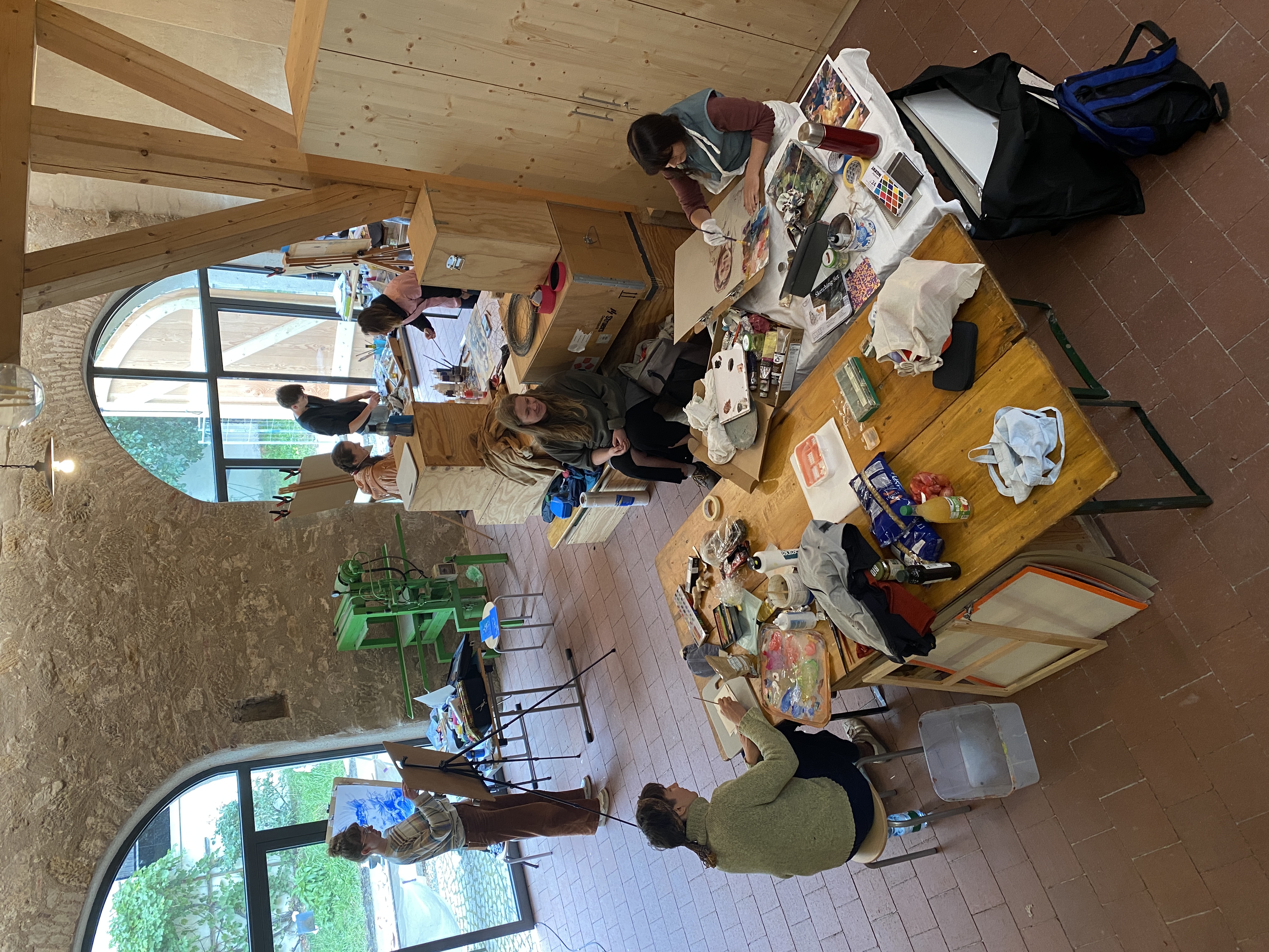 Ein heller, großzügiger Raum mit hohen Decken und großen Fenstern, in dem mehrere Personen an Tischen künstlerisch arbeiten. Auf den Tischen liegen Materialien wie Papier, Farben und Werkzeuge. Einige Personen sind konzentriert bei der Arbeit, andere unterhalten sich oder schauen sich um. Im Hintergrund malt jemand an einer Staffelei. Der Raum vermittelt eine kreative und offene Atmosphäre. (öffnet Vergrößerung des Bildes)