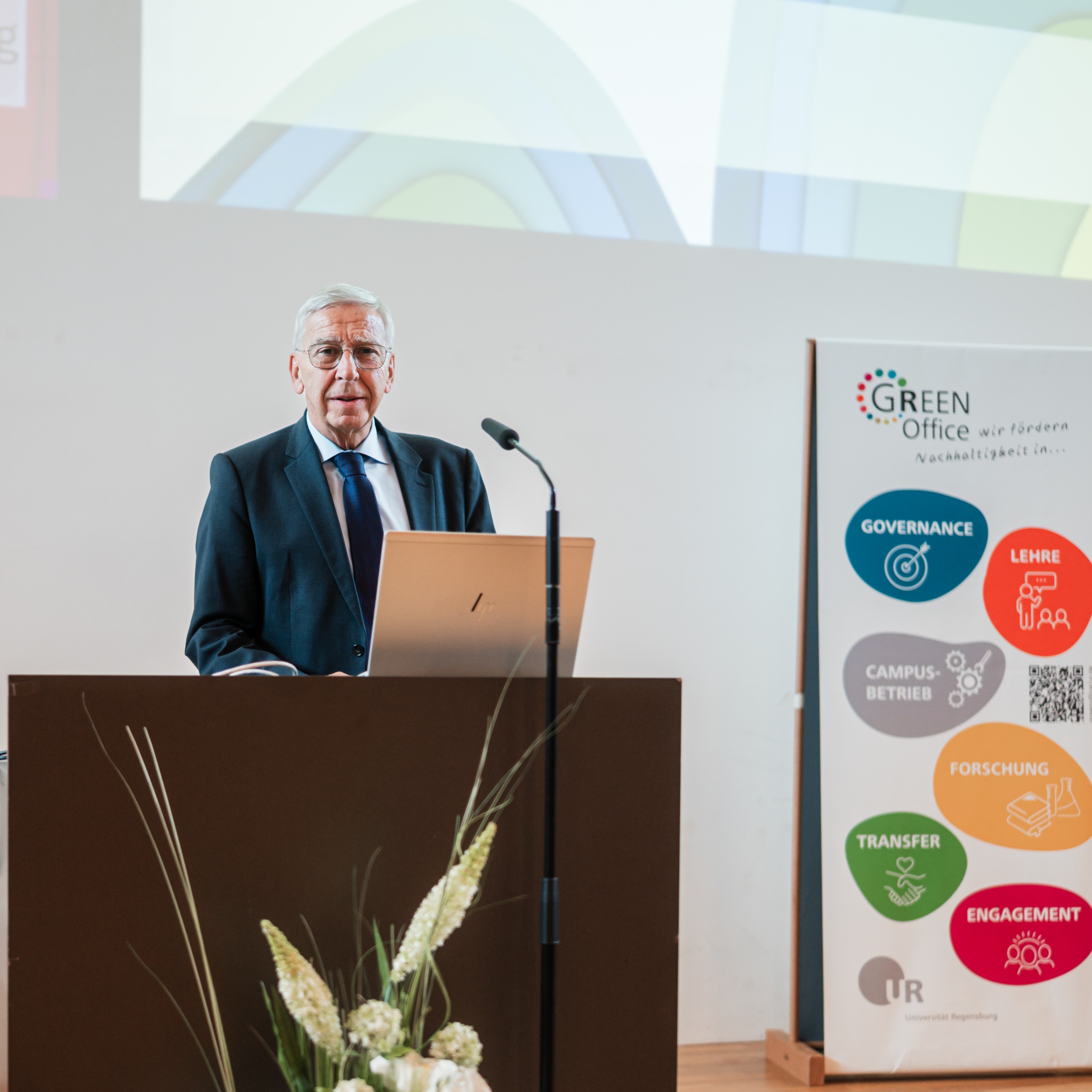 Ein Mann mit grauen Haaren, einem Anzug und einer Brille steht an einem Rednerpult. Im Hintergrund steht ein Aufsteller mit der Aufschrift "Green Office". (öffnet Vergrößerung des Bildes)