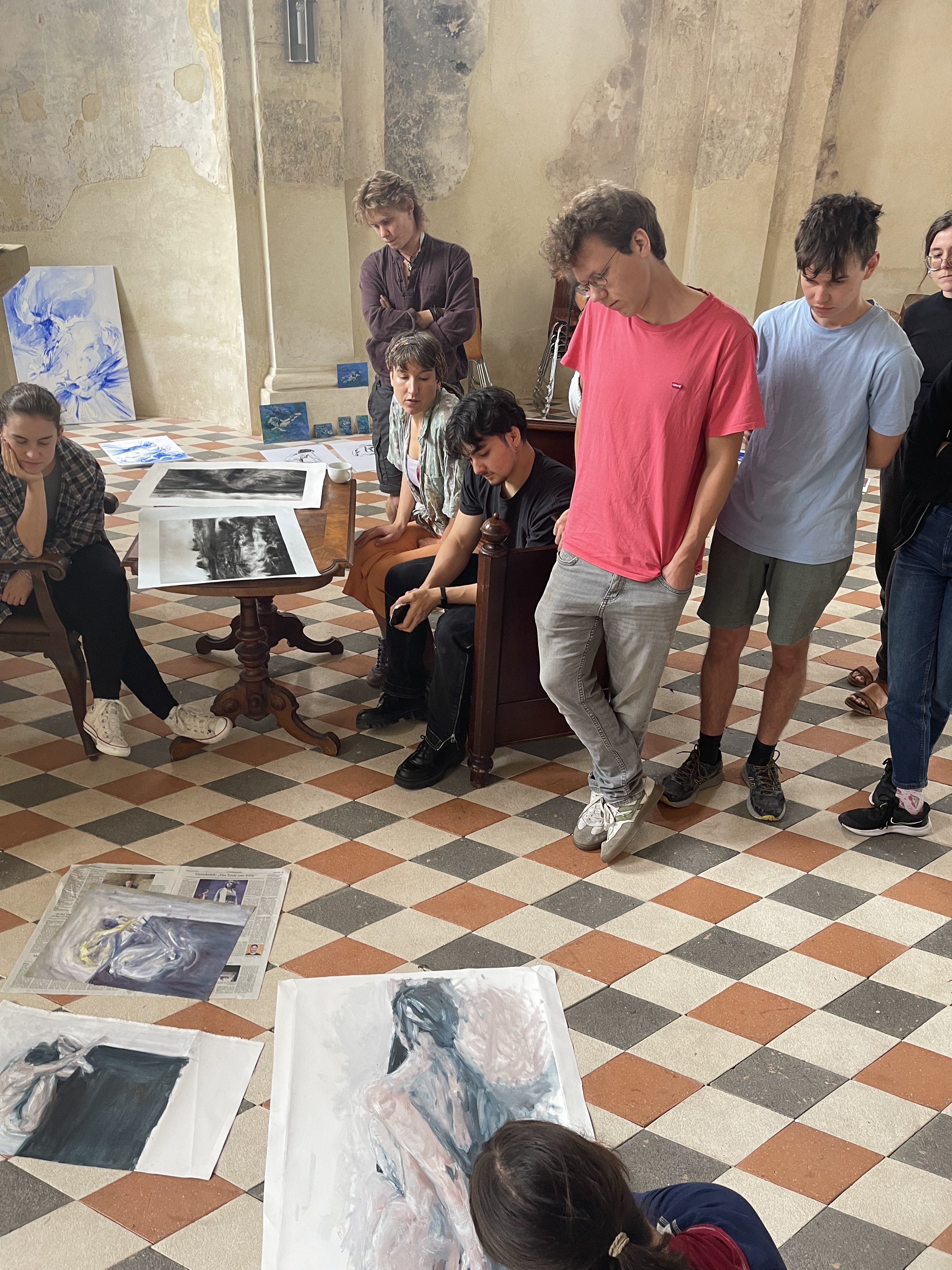 Mehrere Personen befinden sich in einem Raum mit gemustertem Boden und abgenutzten Wänden. Sie stehen oder sitzen um einen Tisch und betrachten verschiedene Kunstwerke, die auf dem Tisch und dem Boden verteilt sind. Die Werke zeigen unterschiedliche Stile und Motive, und die Personen wirken in Gespräche vertieft. (öffnet Vergrößerung des Bildes)