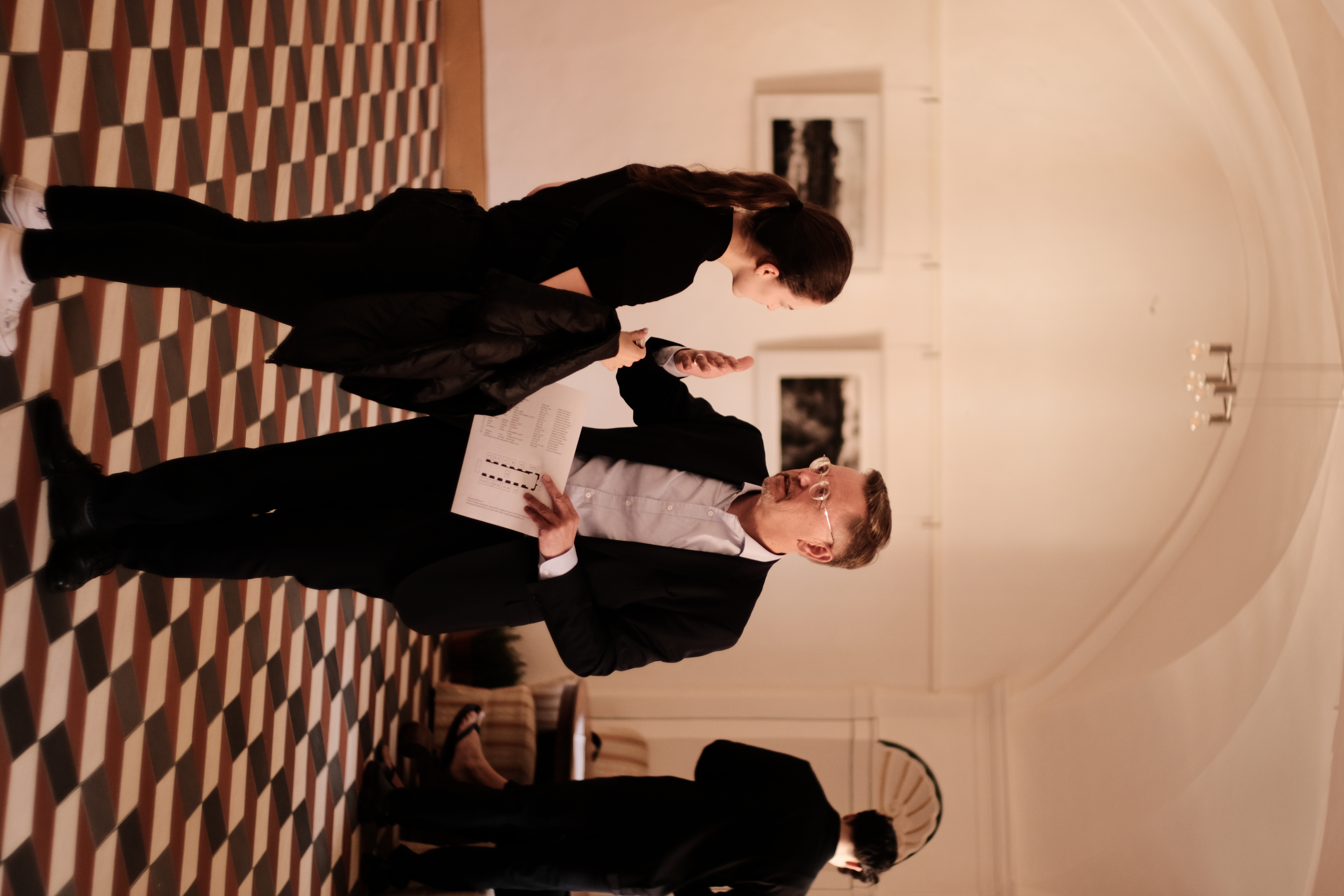 Ein Mann im Anzug spricht mit einer Frau in legerer Kleidung in einem elegant dekorierten Raum mit schwarz-weiß kariertem Boden. Er hält ein Blatt Papier in der Hand, während sie aufmerksam zuhört. Im Hintergrund unterhalten sich zwei weitere Personen vor gerahmten Fotografien an der Wand. (öffnet Vergrößerung des Bildes)