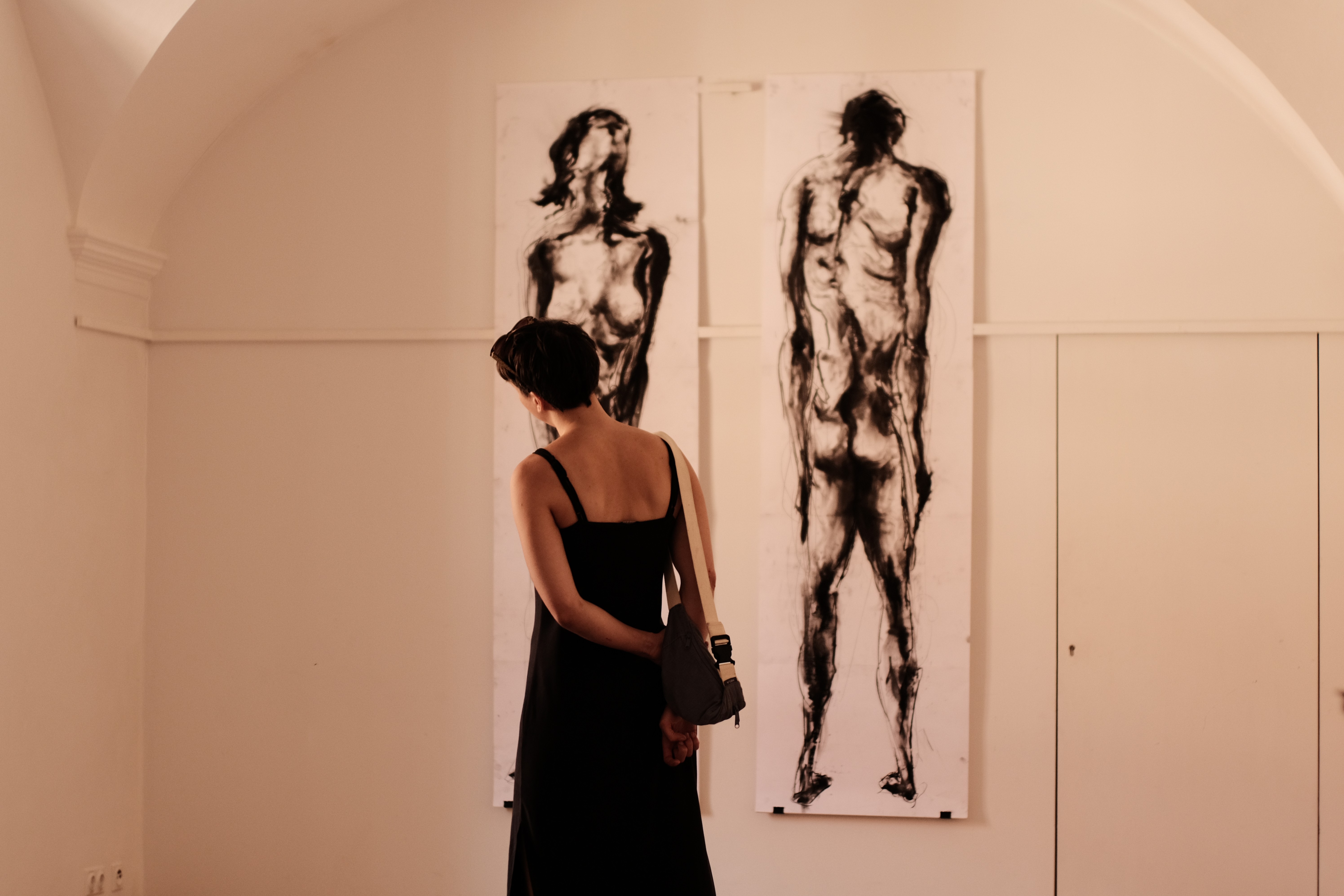 Eine Frau in einem schwarzen Kleid steht mit dem Rücken zur Kamera und betrachtet zwei großformatige, abstrakte Schwarz-Weiß-Kunstwerke an einer weißen Wand. Über ihrer rechten Schulter trägt sie eine schwarze Tasche. (öffnet Vergrößerung des Bildes)
