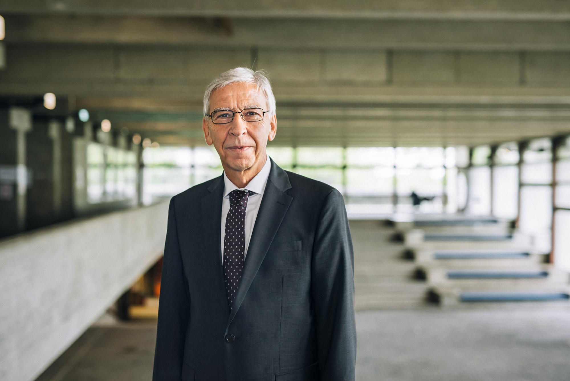 Ein Mann im Anzug und mit Krawatte, mit grauem Kurzhaarschnitt und Brille, steht entspannt und dem Betrachter zugewandt in einem weitläufigen Gebäude mit vielen Fensterfronten.