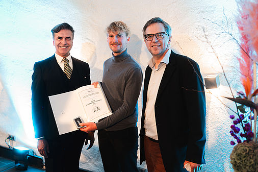 Drei Männer stehen vor einer weißen Wand, rechts im Hintergrund ein buntes Blumengesteck. Der in der Mitte stehende junge Mann trägt einen dunkelgrauen Rollkragenpullover. Er hält dem Betrachter den Inhalt einer aufgeschlagene Urkundenmappe entgegen.