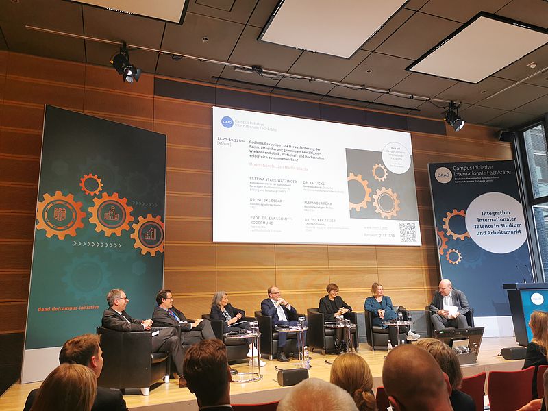 Podiumsdiskussion. Mehrere Personen sitzen in einer Podiumsrunde zusammen vor Publikum.