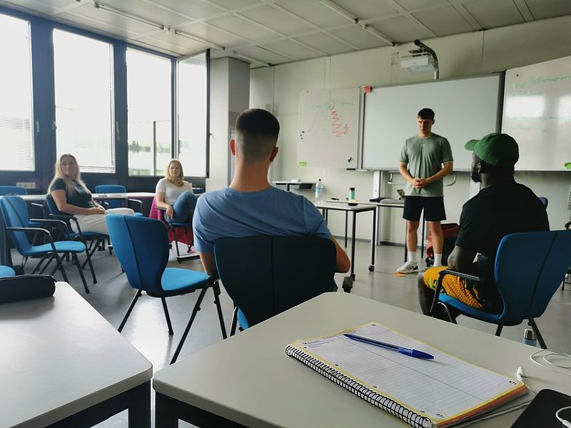 Studierende sitzen in einem Seminarraum und sprechen miteinander. Ein Student steht vorne, die Anderen sitzen in einem Halbkreis vor ihm.