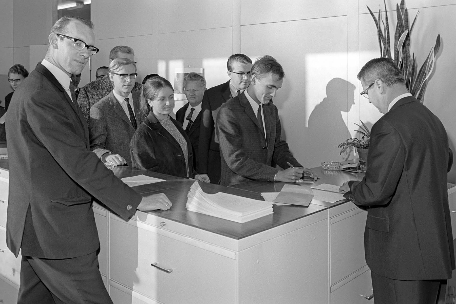 The black and white photograph shows a counter behind which men in suits are standing. On the counter are documents, an ashtray, and potted plants. Behind the counter is a line of young people in suits and dresses. (opens enlarged image)