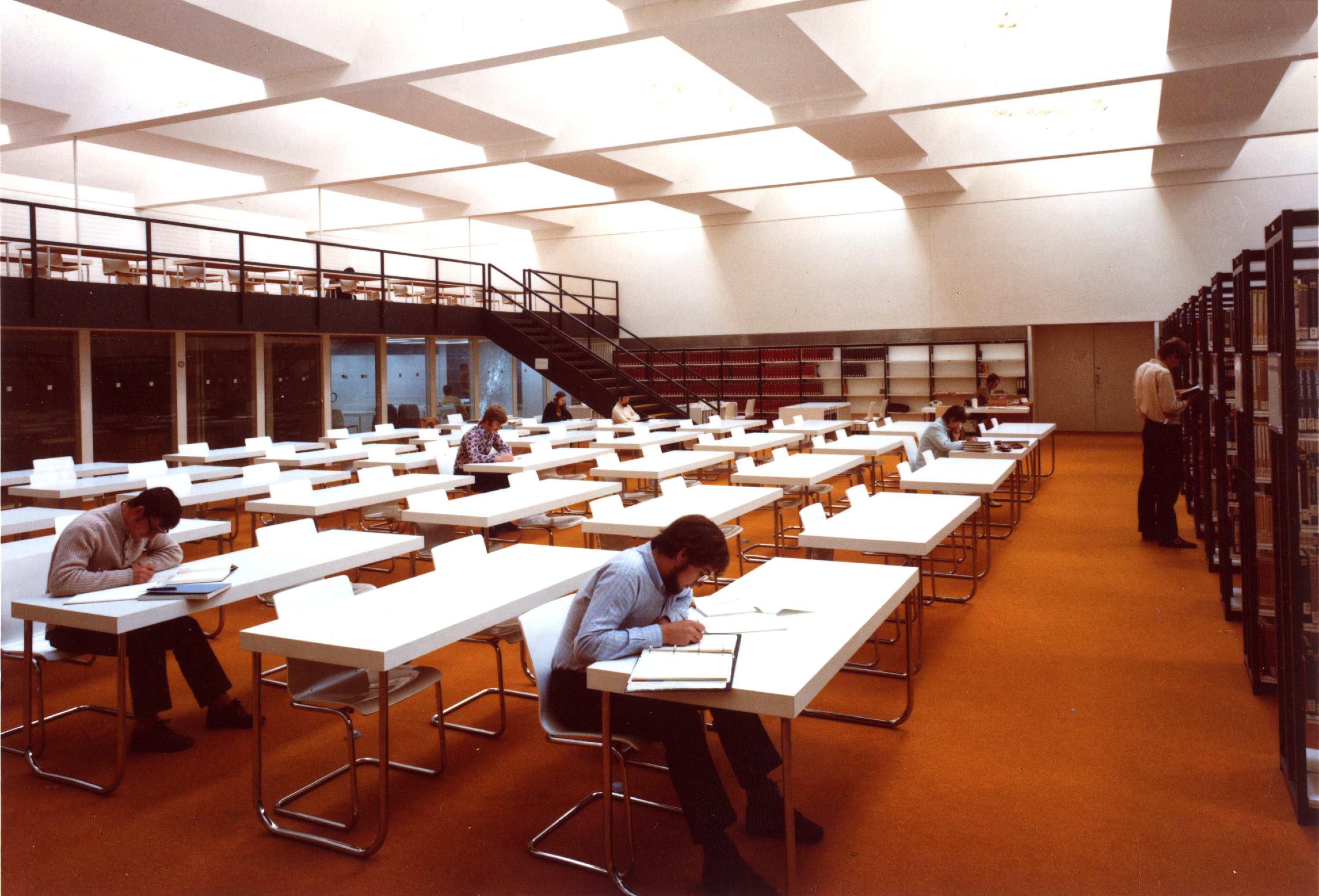 In a reading room with orange carpeting, there are white tables and chairs, and bookshelves line the walls. Young people are sitting at some of the tables, reading or writing. The color scheme of the photograph suggests it was taken in the 1970s. (opens enlarged image)